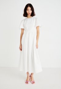 Witte midi-jurk met korte vlindermouwen, voorzien van geperforeerde patronen. Gecombineerd met roze haksandalen met bloemaccenten.