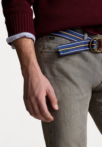 Burgunderröd bomullströja med ribbade manschetter, randig skjorta under, grå denimjeans och ett blått bälte med gula ränder och en metallspänne.