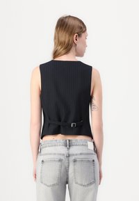 Schwarzes ärmelloses Top mit Nadelstreifen und einem rückseitigen Schnallendetail, kombiniert mit hellgrauen Jeans mit Gesäßtaschen. Glatter Stofftextur.