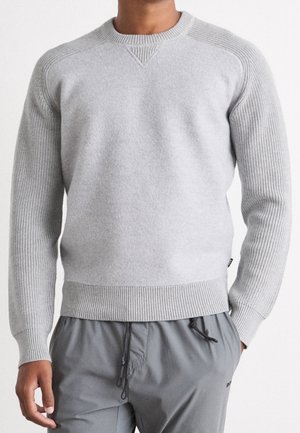 Mann trägt einen hellgrauen Strickpullover mit gerippten Ärmeln und Saum, kombiniert mit grauen Kordelhosen, stehend vor weißem Hintergrund.