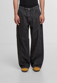 Loose-fit black denim jeans with visible stitching, a high waist, and curved side panels, paired with yellow sneakers.