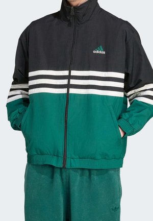 Mężczyzna noszący kurtkę zapinaną na zamek Adidas z czarnym wierzchem, białymi paskami i zielonym dołem, ręce w kieszeniach, w połączeniu z zielonymi spodniami.