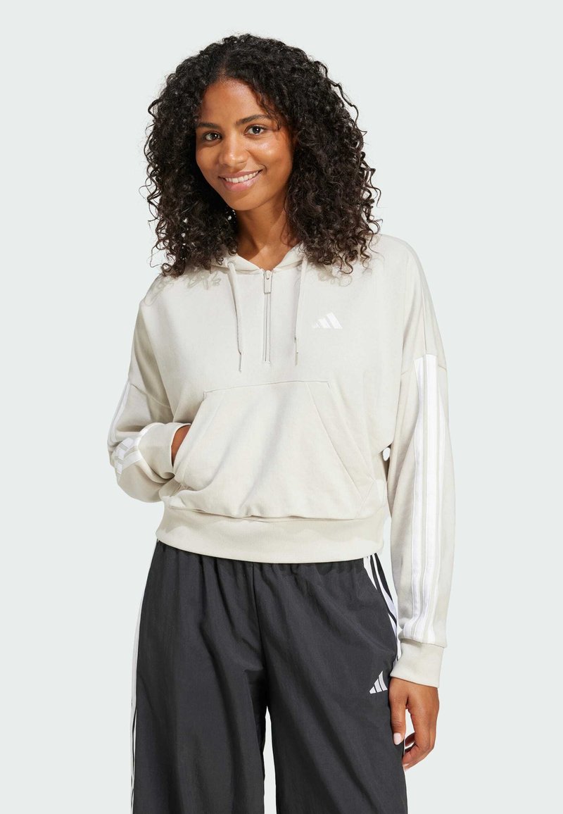 Beige hooded sweatshirt with a front pocket, half zip, and white stripe accents on sleeves. Paired with loose black pants featuring white stripes.