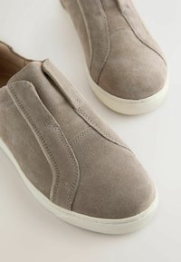 Baskets slip-on en daim beige avec semelles en caoutchouc blanches, coutures visibles et bouts arrondis lisses sur un fond clair.