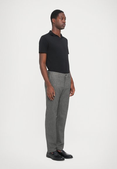 Schwarzes Polo-Shirt mit kurzen Ärmeln, graue strukturierte Anzughose und schwarze Slipper. Das Model steht im Profil vor einem weißen Hintergrund.