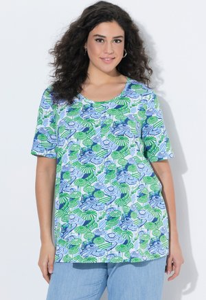 Uśmiechnięta kobieta o kręconych włosach, nosząca srebrne kolczyki w kształcie koła, niebiesko-zieloną koszulę z krótkim rękawem w wzory oraz jasnoniebieskie spodnie.