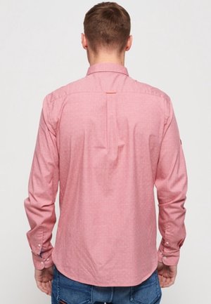 Mand iført en pink langærmet skjorte med knapper og blå jeans, vist bagfra mod en ensfarvet baggrund.