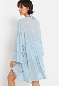 Vestido de cobertura transparente azul claro, con mangas anchas y un lazo ajustable en la parte posterior. Presenta una forma acampanada y un acabado texturizado.