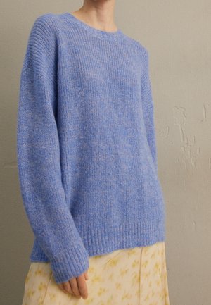 Personne portant un pull léger bleu clair en maille douce et ample, et une jupe jaune pâle avec un motif floral subtil, debout contre un mur uni.