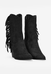 Botas de tobillo de ante negro con punta afilada y tacón alto, que cuentan con detalles de flecos en los laterales y una cremallera en el tobillo interno.