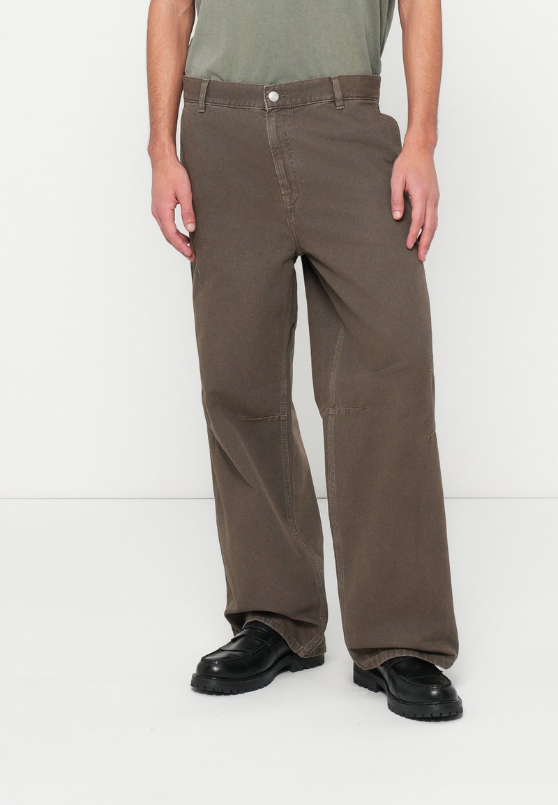 Pantalones de pierna ancha marrones de algodón con una textura lisa, con cierre frontal de botón y bolsillos laterales, llevados con zapatos negros.