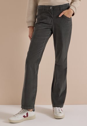 Personne portant un pantalon droit gris foncé, un haut beige à manches longues et des baskets blanches avec des accents rouges, debout avec une main dans la poche.