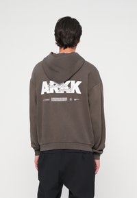 Barna kapucnis pulóver, laza szabással, hátul fehér grafikával és bordázott mandzsettákkal, valamint alsó szegéllyel. Puha anyagú textúra.