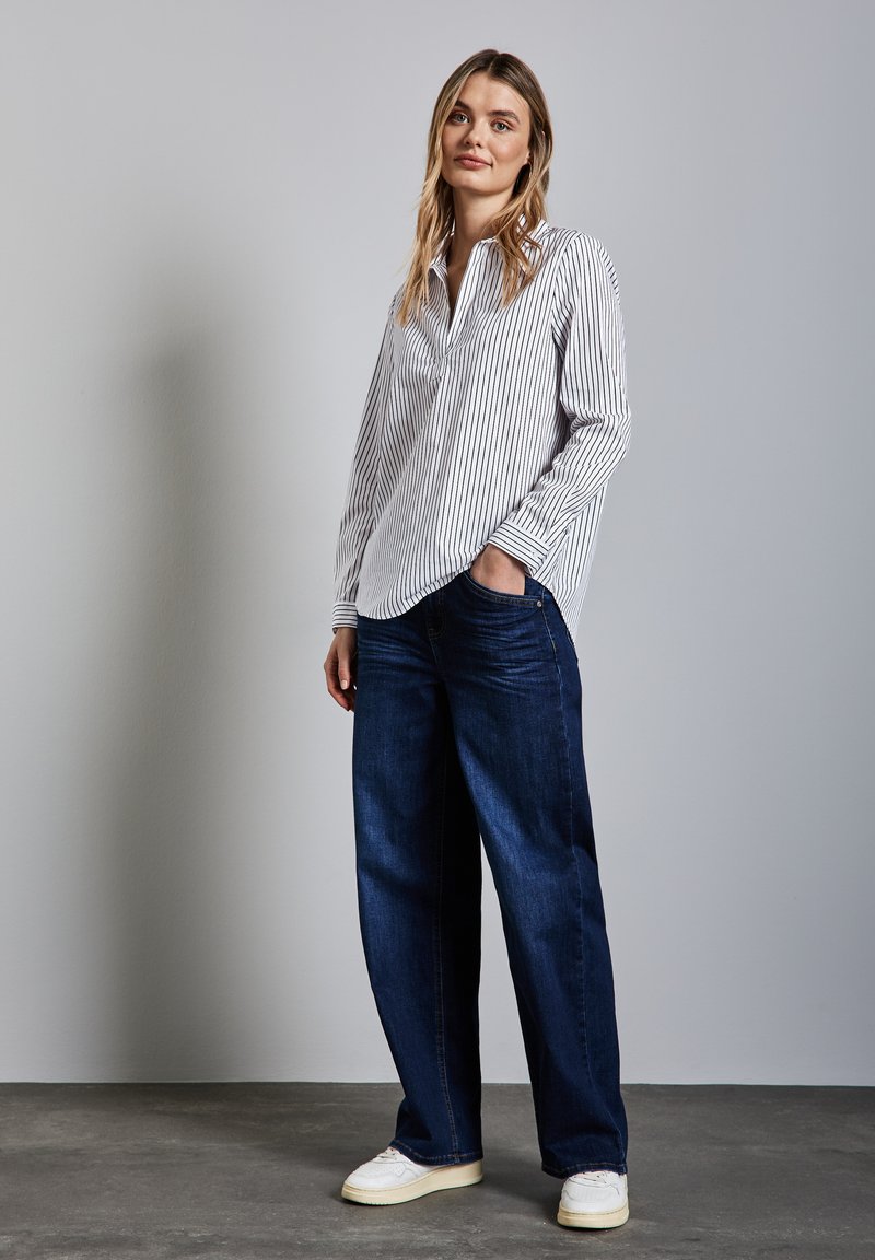 Randig vit- och marinblå skjorta med knappar, kombinerad med vida mörka denimjeans och vita sneakers, stående på en grå yta mot en neutral bakgrund.