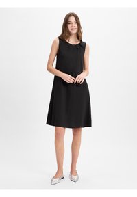 Zwarte mouwloze jurk met een ronde kraag, gemaakt van lichte stof, valt tot halverwege de dij en is gecombineerd met zilveren puntige ballerina's.