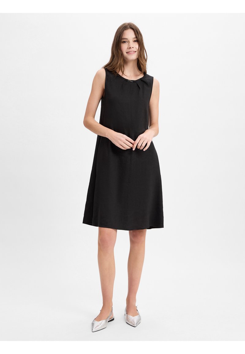 Zwarte mouwloze jurk met een ronde kraag, gemaakt van lichte stof, valt tot halverwege de dij en is gecombineerd met zilveren puntige ballerina's.