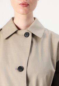 Un pardesiu bej cu un colier clasic, având un nasture negru, o textură fină și o croială ușor ajustată, punând accent pe linii curate.