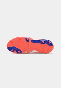 Zapato de fútbol con una suela vibrante en naranja y azul, parte superior blanca, agarre texturizado y clavos prominentes para tracción.