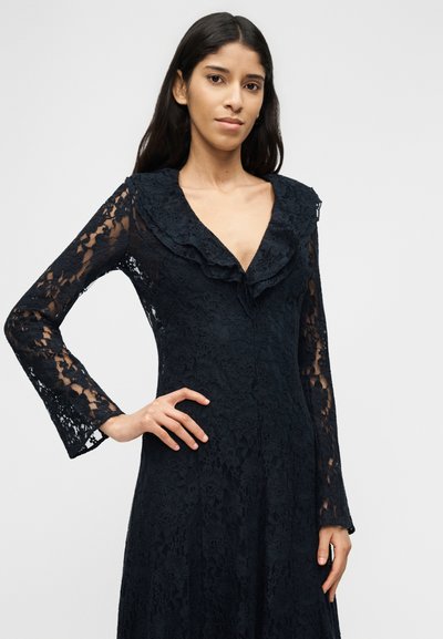Γυναίκα με μακριά σκούρα μαλλιά που φοράει μαύρο φόρεμα από δαντέλα με βολάν στο γιακά και μακριά μανίκια, ποζάροντας με το ένα χέρι στη μέση.