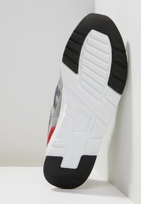 Basket gris avec une tige texturée, dotée d'une semelle en caoutchouc noire et rouge. Un logo proéminent et des éléments de design renforcent son attrait visuel.