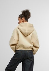 Beige oversized mikina s kapucňou na šnúrku, rebrovanými okrajmi a spadnutými ramenami, v kombinácii s tmavými džínsami s vreckami na zadočku a šitými detailmi.