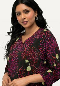 Blusa nera con un vivace motivo floreale in viola e verde, scollatura a V, maniche lunghe e una texture morbida. Orecchini in argento visibili.