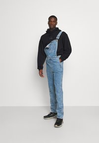 Τζιν φόρμες σε ανοιχτό μπλε, διαθέτοντας ένα μόνο λουρί στον ώμο, ίσια πόδια και μπροστινή τσέπη. Φορεμένες με μια μαύρη φούτερ και μαύρα αθλητικά παπούτσια.