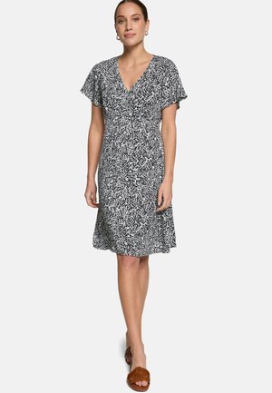 Žena v čierno-bielych šatách po kolená s motívom listov, krátkymi rukávmi a hnedými sandálmi s otvorenou špičkou, stojaca pred jednoduchým pozadím.