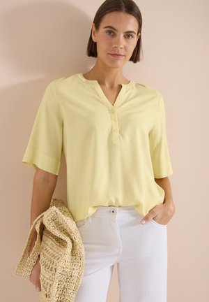 Mujer con blusa de manga corta de color amarillo claro y pantalones blancos, sosteniendo un bolso tejido color beige, de pie contra una pared lisa.