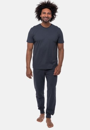 Smilende mand med krøllet hår, der går barfodet, iført en mørkegrå kortærmet skjorte og matchende joggingbukser mod en ensartet lys baggrund.