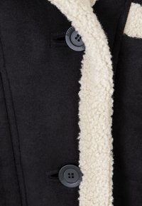 Czarny płaszcz z kremowym, puszystym kołnierzem i dwoma czarnymi guzikami. Tkanina wydaje się być miękka, ma gładką teksturę i zdefiniowany fason.