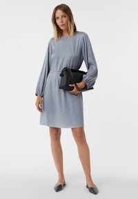 Jasnoszara sukienka z długim rękawem, z talią ściągniętą, gładką fakturą i luźnym krojem. W zestawie z czarną torebką i butami na szpilkach.