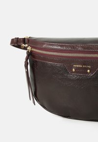 Bordeaux leren crossbody tas met gouden hardware, een gebogen vorm, een sluiting met een rits aan de bovenkant en een reliëflogo op de voorkant.