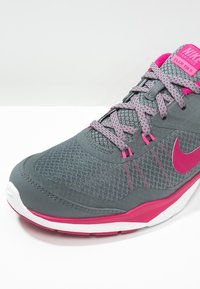 Zapatilla de entrenamiento Nike con parte superior gris, acentos rosas, textura de malla y suela blanca. Presenta cordones y una marca en la lengua.