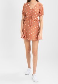 Kurzes V-Ausschnitt-Kleid in rostorange mit weißen Punkten; mit einem Kordelzug in der Taille und einem ausgestellten Saum. Getragen mit weißen Sneakers.