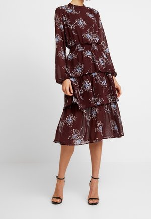 Vrouw draagt een bordeauxrode midi-jurk met bloemenprint, lange mouwen, een gelaagde rok en zwarte enkellaarzen met bandje, staand voor een eenvoudige lichte achtergrond.