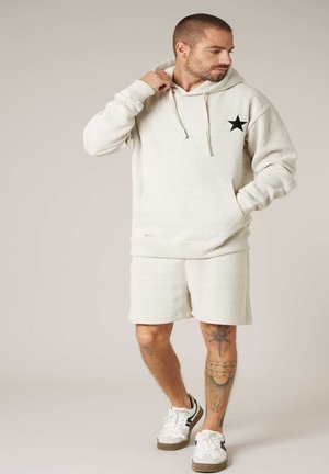 Homme portant un sweat à capuche crème avec une étoile noire, un short assorti et des baskets blanches, debout et regardant sur le côté avec la main sur la capuche.