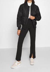 Černá bomber bunda s žebrovanými manžetami a límcem, doplněná černými kalhotami s vysokým pasem a postranními rozparky, a neutrálními teniskami.