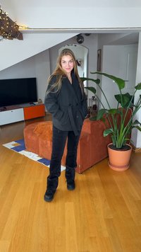 Svart rock med bälte i midjan, kombinerad med svarta byxor, placerad i en modern interiör med trägolv och en grön växt.