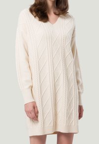 Cremefarbenes gestricktes Kleid mit V-Ausschnitt, das ein strukturiertes Rauten-Muster und gerippte Bündchen aufweist und oberhalb des Knies endet.