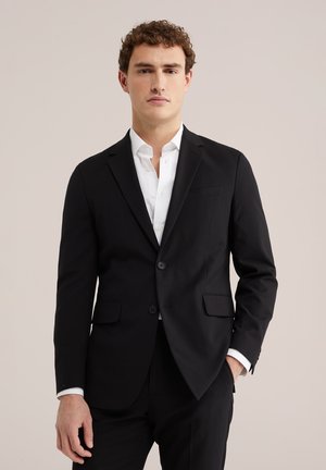 Joven vestido con un traje negro con chaqueta abotonada y una camisa blanca, posando frente a un fondo claro y liso.