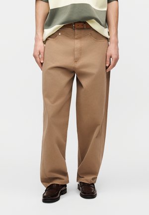 Személy laza szabású bézs nadrágban, barna övvel és sötétbarna bőr cipőben, sima világos háttér előtt állva.