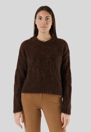 Donna con lunghi capelli scuri che indossa un maglione marrone scuro testurizzato e pantaloni marrone chiaro, in piedi davanti a uno sfondo grigio semplice.