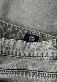Țesătură de denim gri cu o suprafață texturată și un model decolorat. Dispune de un buton negru și o piesă de talie. Detalii de cusut vizibile.