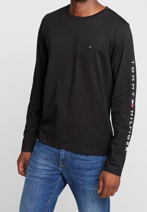 Mężczyzna w czarnej bluzce z długim rękawem z napisem "Tommy Hilfiger" i logo na prawym rękawie, w niebieskich jeansach, na jednolitym tle.