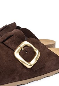 Braune Wildleder-Slidesandalen mit einer großen goldenen Schnalle, ausgestattet mit einem konturierten Fußbett und einer strukturierten Laufsohle. Weiches, unauffälliges Design.