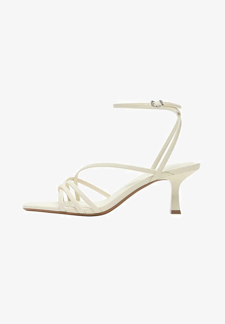Sandale albă cu toc înalt, cu barete subțiri, o baretă cu cataramă la gleznă și un toc subțire de înălțime medie, pe fundal alb.