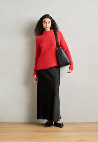 Roter gerippter Strickpullover, schwarze Satin-Maxirüsch, schwarze Umhängetasche und schwarze flache Schuhe. Hintergrund grau, Boden Holz.