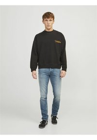 Sweat-shirt noir avec des poignets côtelés, arborant un logo jaune sur la poitrine gauche. À associer avec un jean bleu clair et des baskets noires.