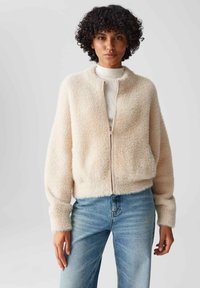 Frau mit lockigem Haar, die eine beigefarbene, flauschige Zip-Jacke über einem hellen Rollkragenpullover und blauen Jeans trägt, vor einem schlichten, hellen Hintergrund steht.